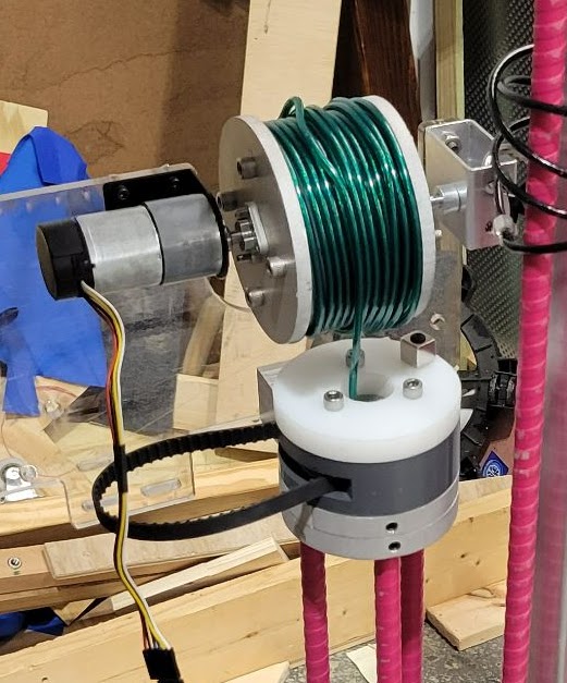 A small motor driving an alumninum spool with coated steel wire wrapped around it. The motor and spool are mounted to a mounting plate, and the wire continues downward through the testing device.